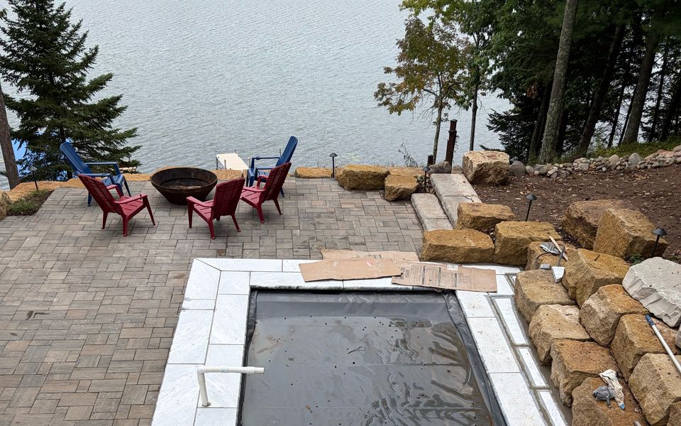 Lakeside patio and fiberglass pool with stone coping and safety cover on Long Lake, Wisconsin, Adirondack chairs and fire pit at the water